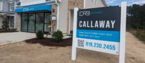 Sales office of a new home community with a large sign displaying visiting hours and contact information.