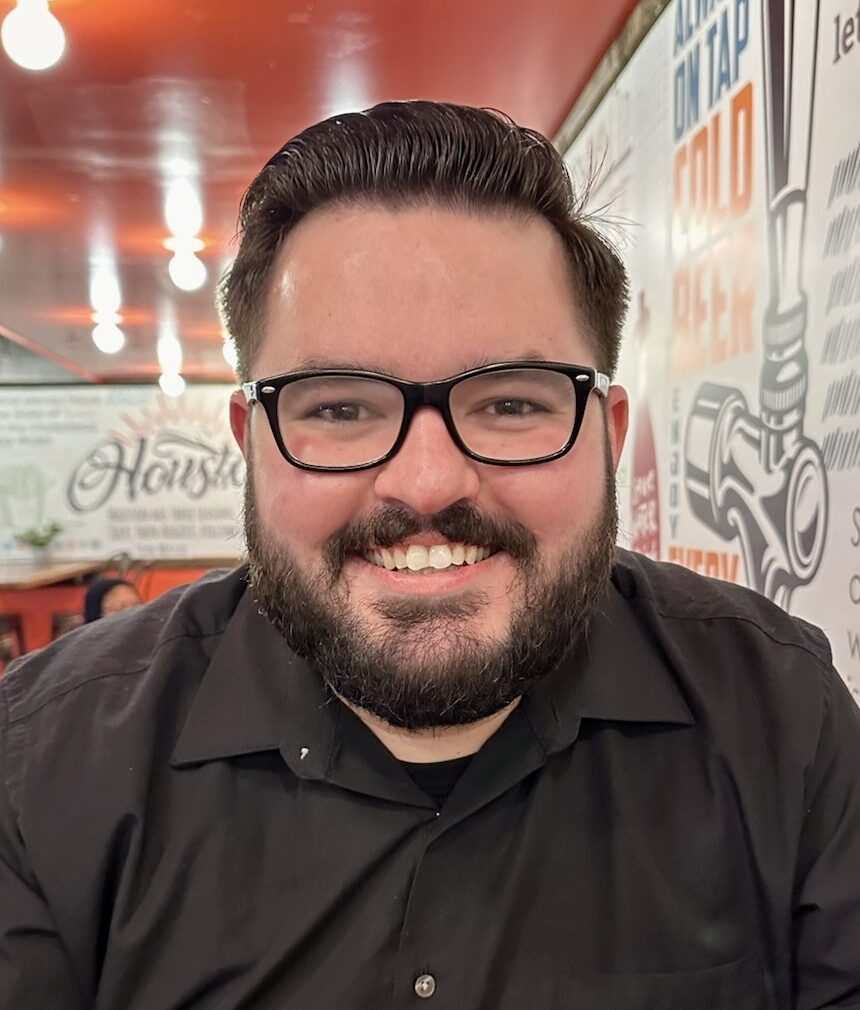 Smiling man with dark hair and beard wearing black shirt and glasses in a retro diner-like restaurant.